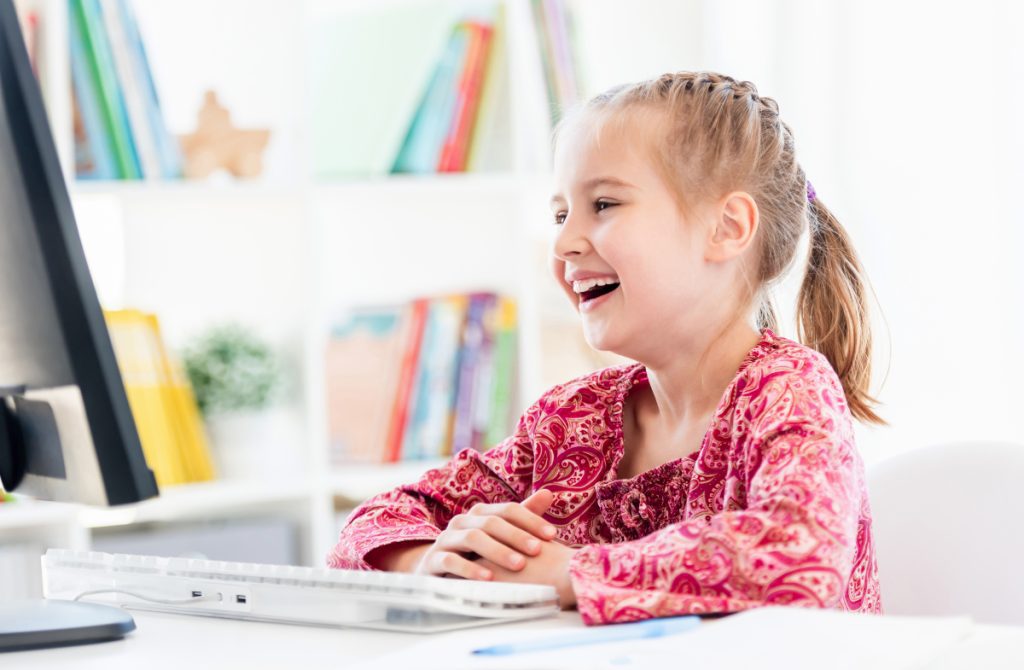 Happy little girl looking at computer screen sitting at desk in classroom